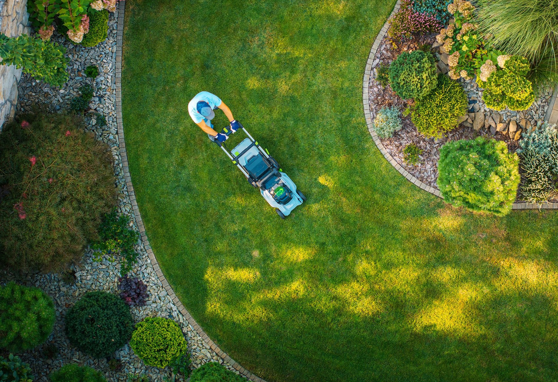 A person mows a lush green lawn in a beautifully landscaped garden with flowerbeds and decorative plants during bright daylight