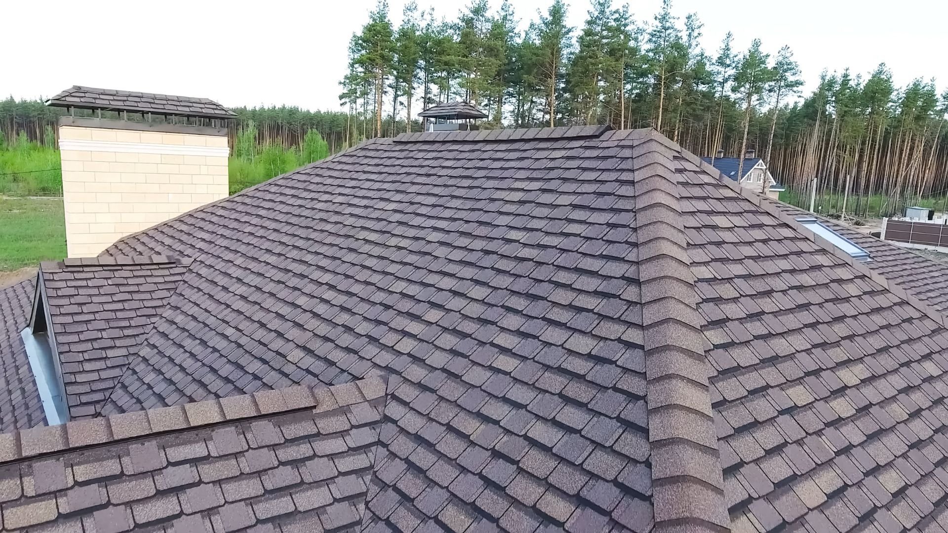 Aerial view of residential roof with dark asphalt shingles, garage and surrounding forest trees