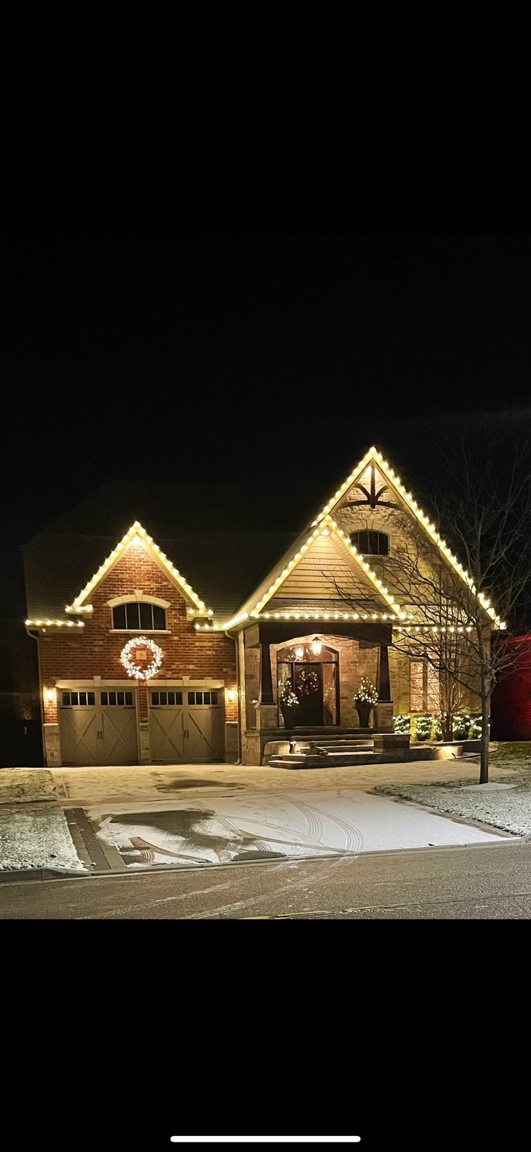 Brick house with decorative holiday lights on roof edges and around entrance at night, wreath on garage door