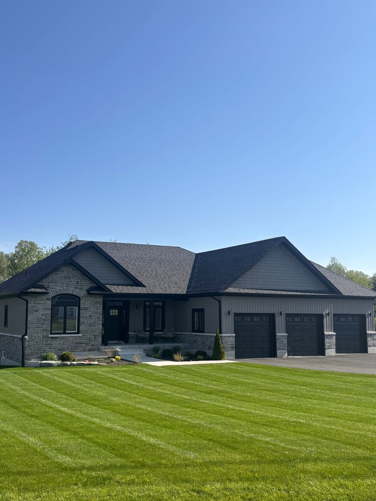 Modern residential house with stone and siding exterior, dark roof, three-car garage, manicured lawn, and clear blue sky