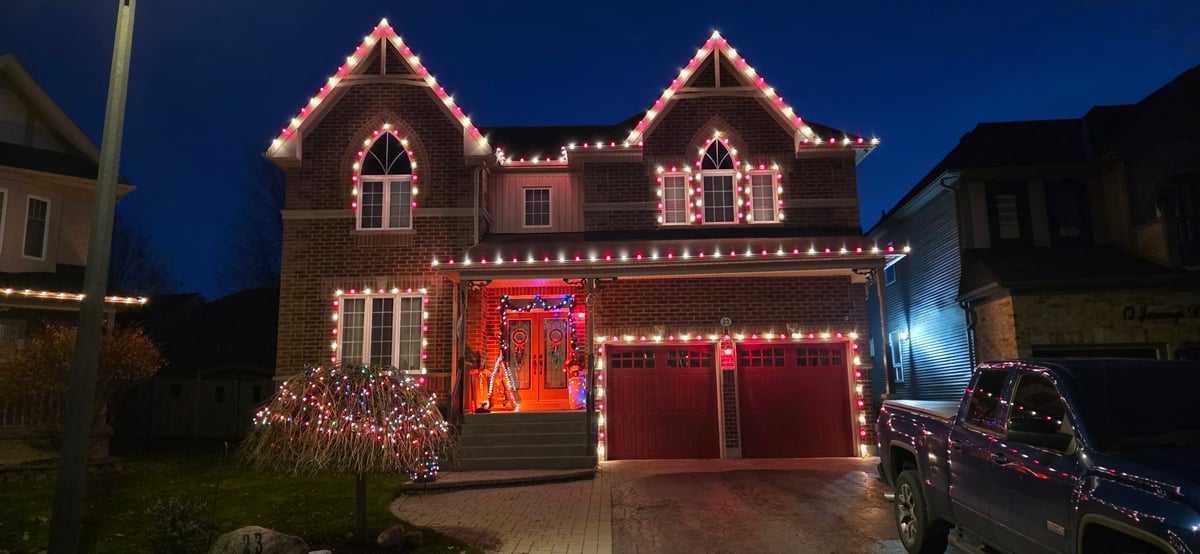 Brick house decorated with red and warm white Christmas lights outlining the roof, windows, and garage doors at dusk